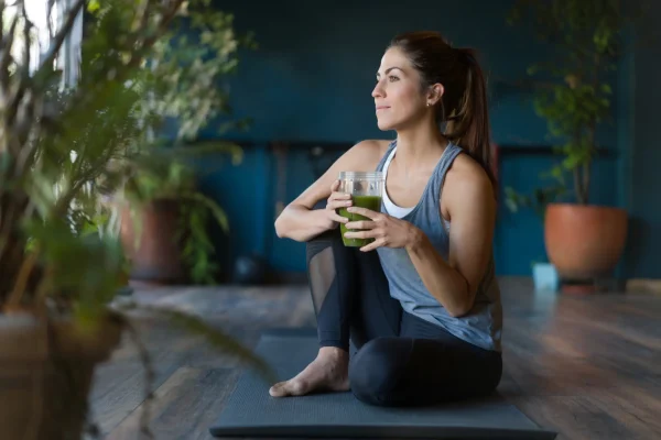 Frau sitzt entspannt auf einer Yogamatte und hält einen grünen Smoothie – Symbol für Gesundheit und private Vorsorge