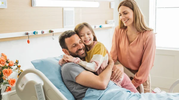 Mann liegt im Krankenhausbett und wird von seiner Tochter und Partnerin umarmt, alle lachen, helle Patientenzimmer-Atmosphäre